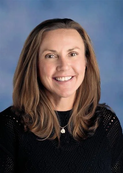 Professional woman with shoulder-length brown hair smiling, wearing a black button-up shirt, against a blue background.
