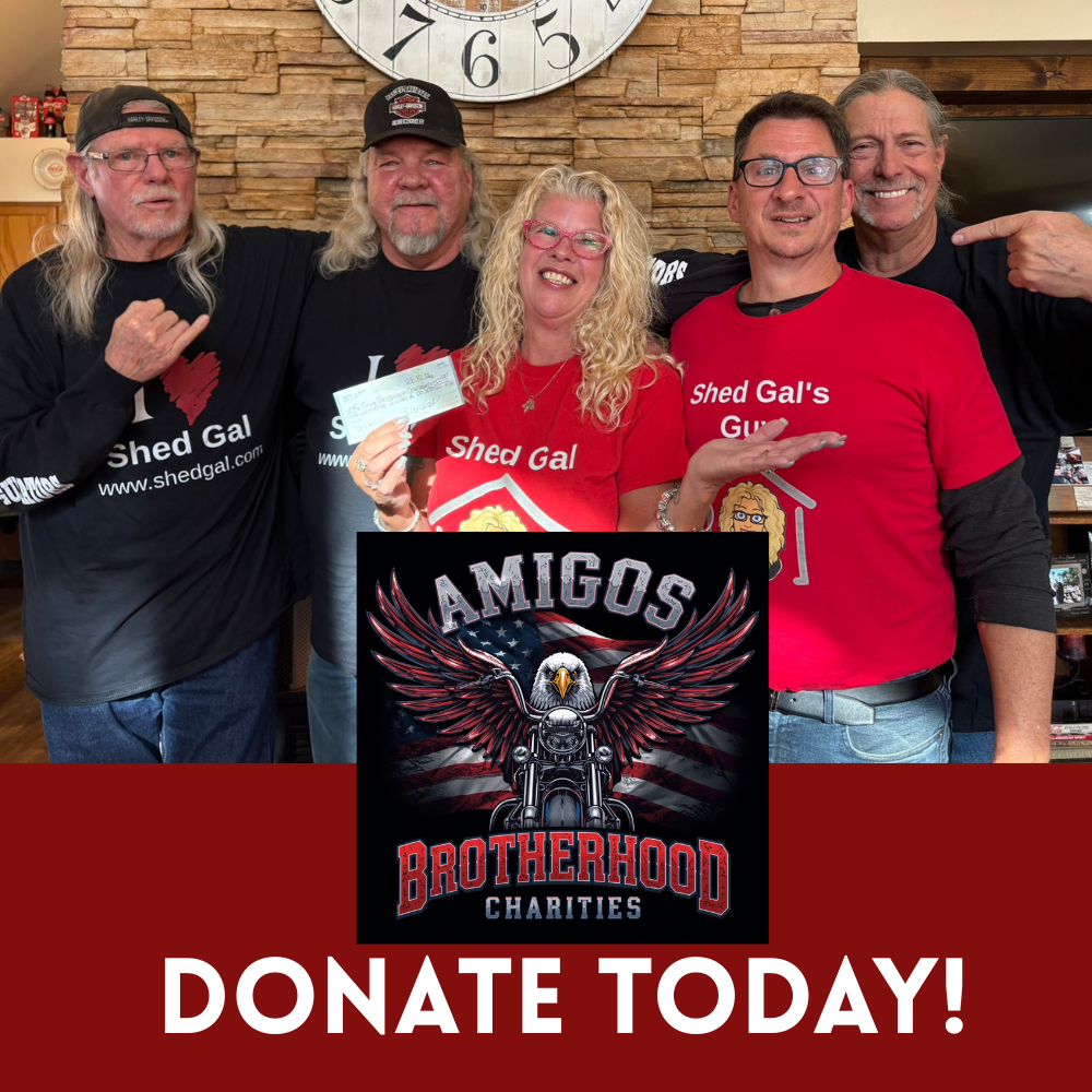 Group of five people standing together, wearing red and black 'Shed Gal' shirts, smiling and posing for a photo in front of a stone wall and large clock. The woman in the center is holding a check, and all are making gestures of support. A charity br