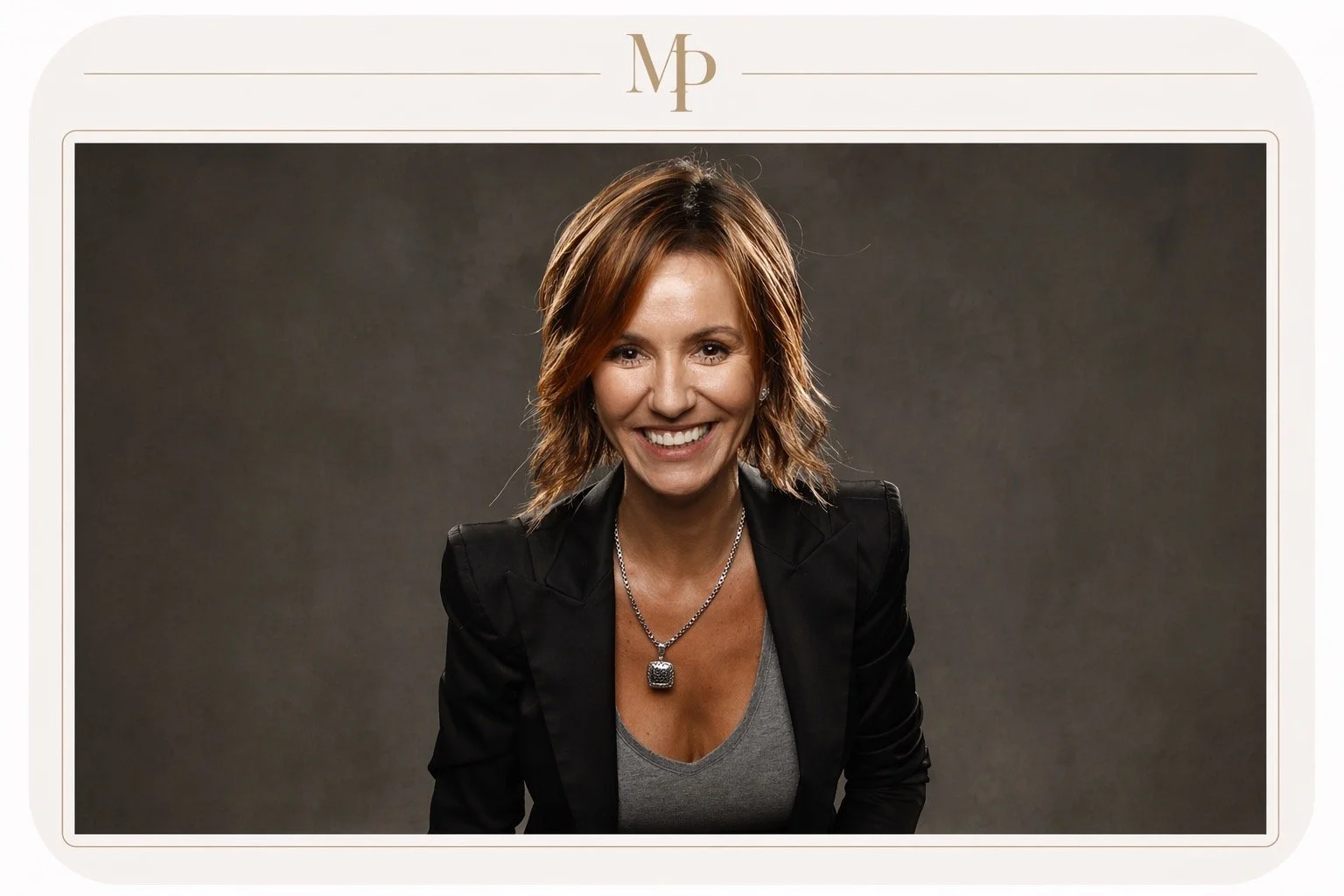 Smiling woman with shoulder-length brown hair wearing a black blazer, gray top, and a silver necklace against a dark gray background.
