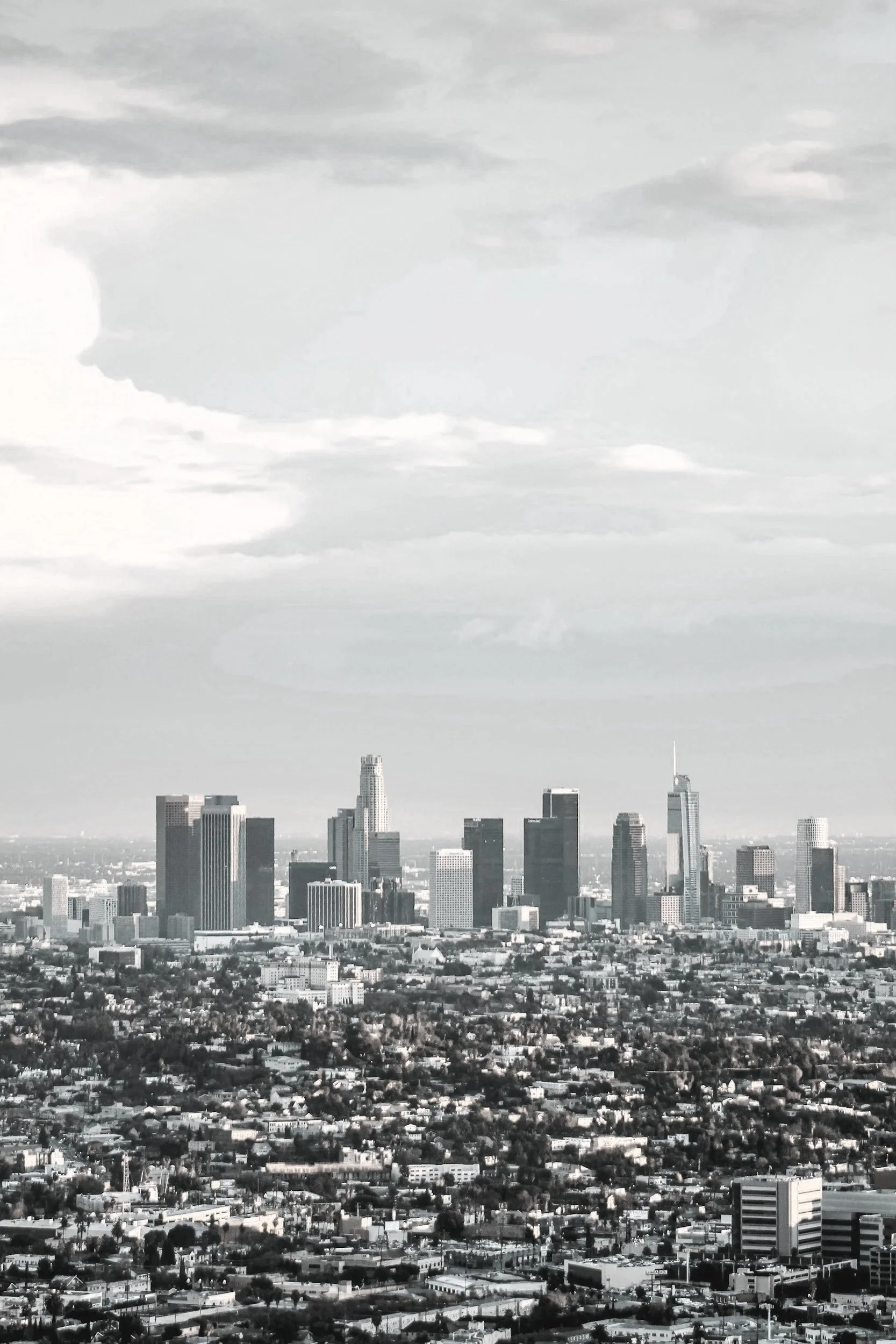 A black and white photo of the Los Angeles skyline with many tall buildings, with a dramatic cloudy sky overhead.