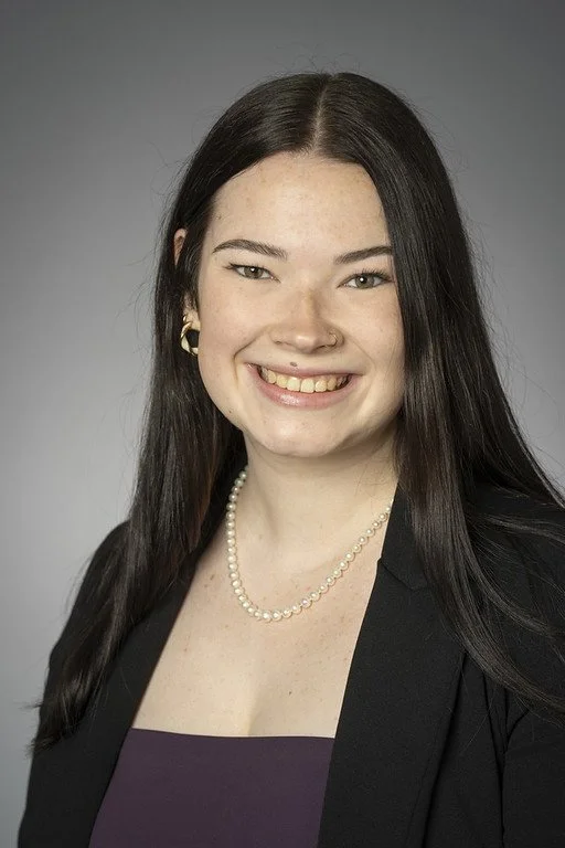 Portrait of a young woman with long dark hair, smiling, wearing a black blazer, purple top, pearl necklace, and gold earrings against a gray background.