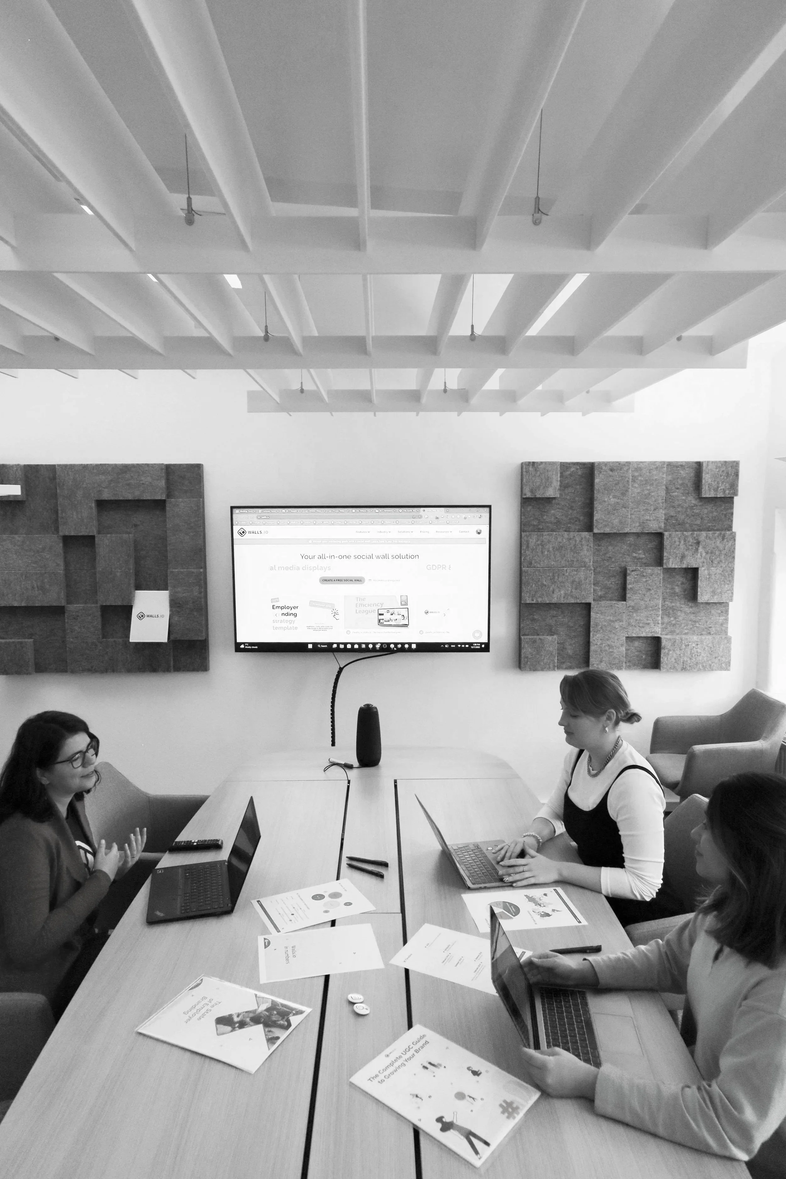 Three women sitting at a conference table in a meeting room with a large screen displaying a presentation. The table has laptops, documents, and pens. The room has acoustic panels on the wall.
