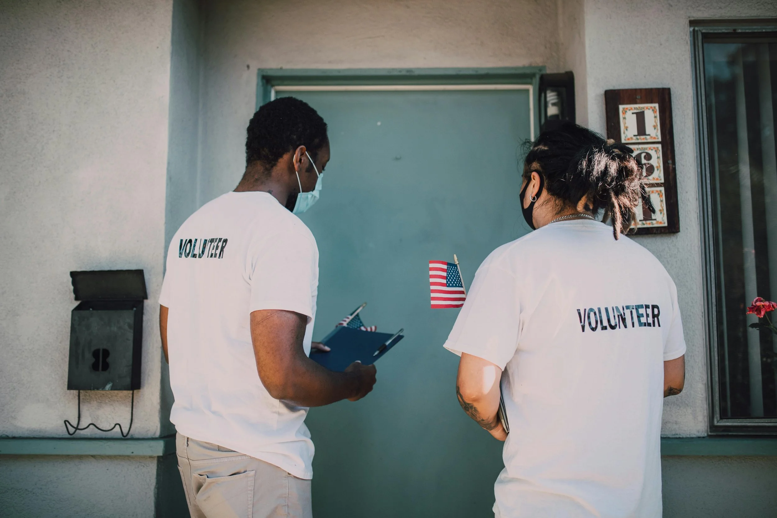 Two volunteers wearing white T-shirts with 'VOLUNTEER' written on the back, standing near a front door. One is holding a clipboard, and the other is holding a pen. The woman has tattoos on her arms, and both are wearing face masks.