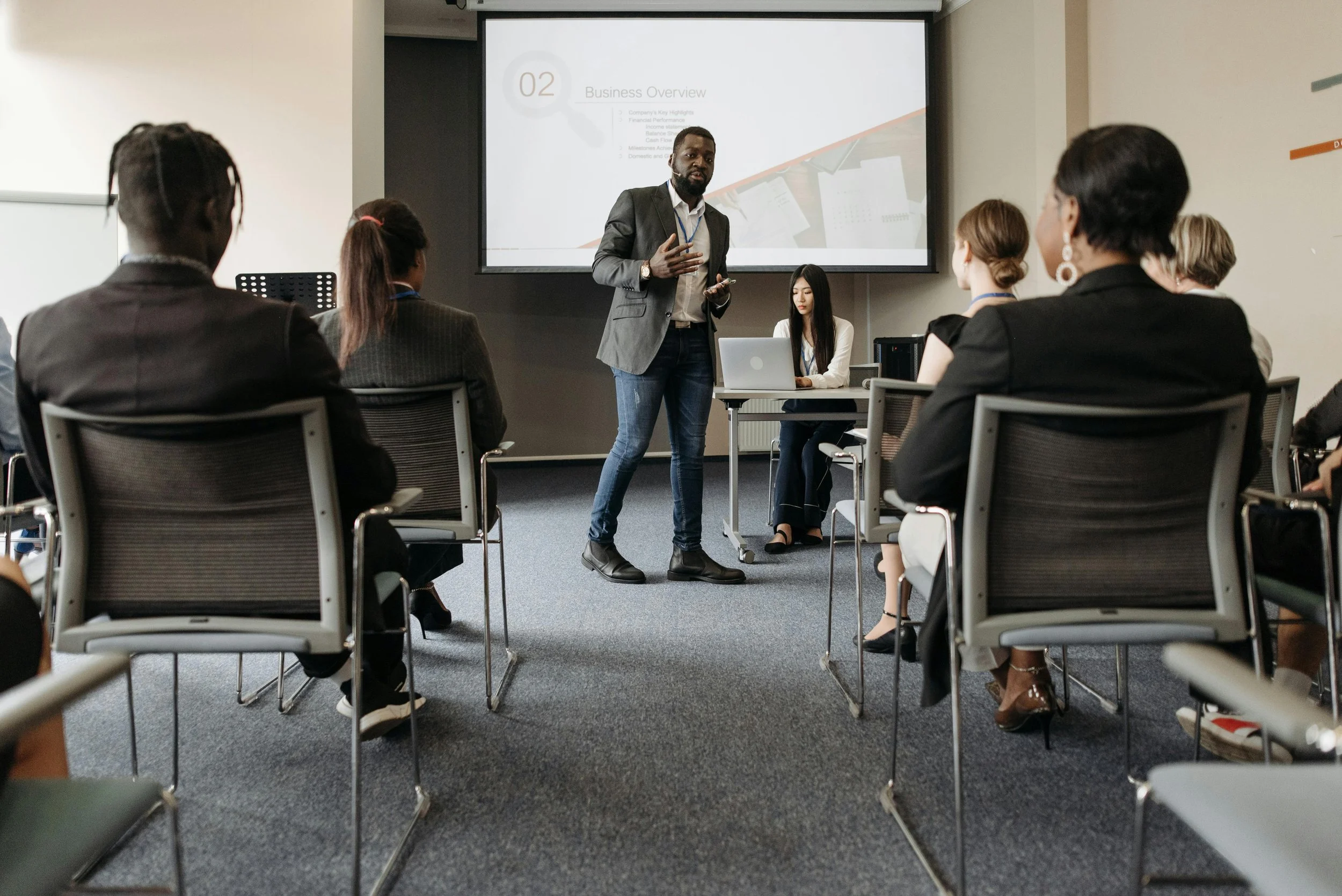 A man giving a business presentation to a group of professionals in a conference room with a large screen displaying a business overview slide.