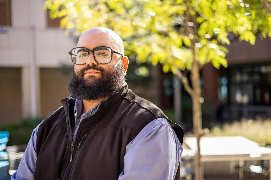 A bald man with a beard and glasses standing outdoors in front of a tree with green leaves, wearing a dark vest and a light blue shirt.
