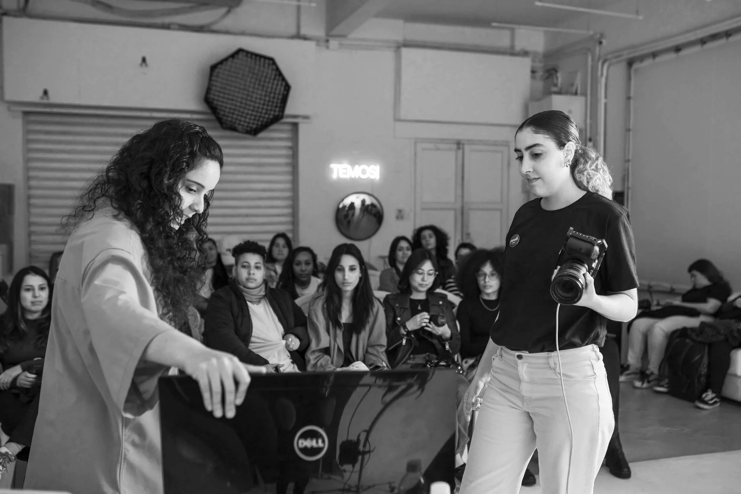 A woman with curly hair is interacting with a Dell computer while a woman with a camera stands nearby, in front of an audience of women in a room with a neon sign that reads 'TEMOSI'.