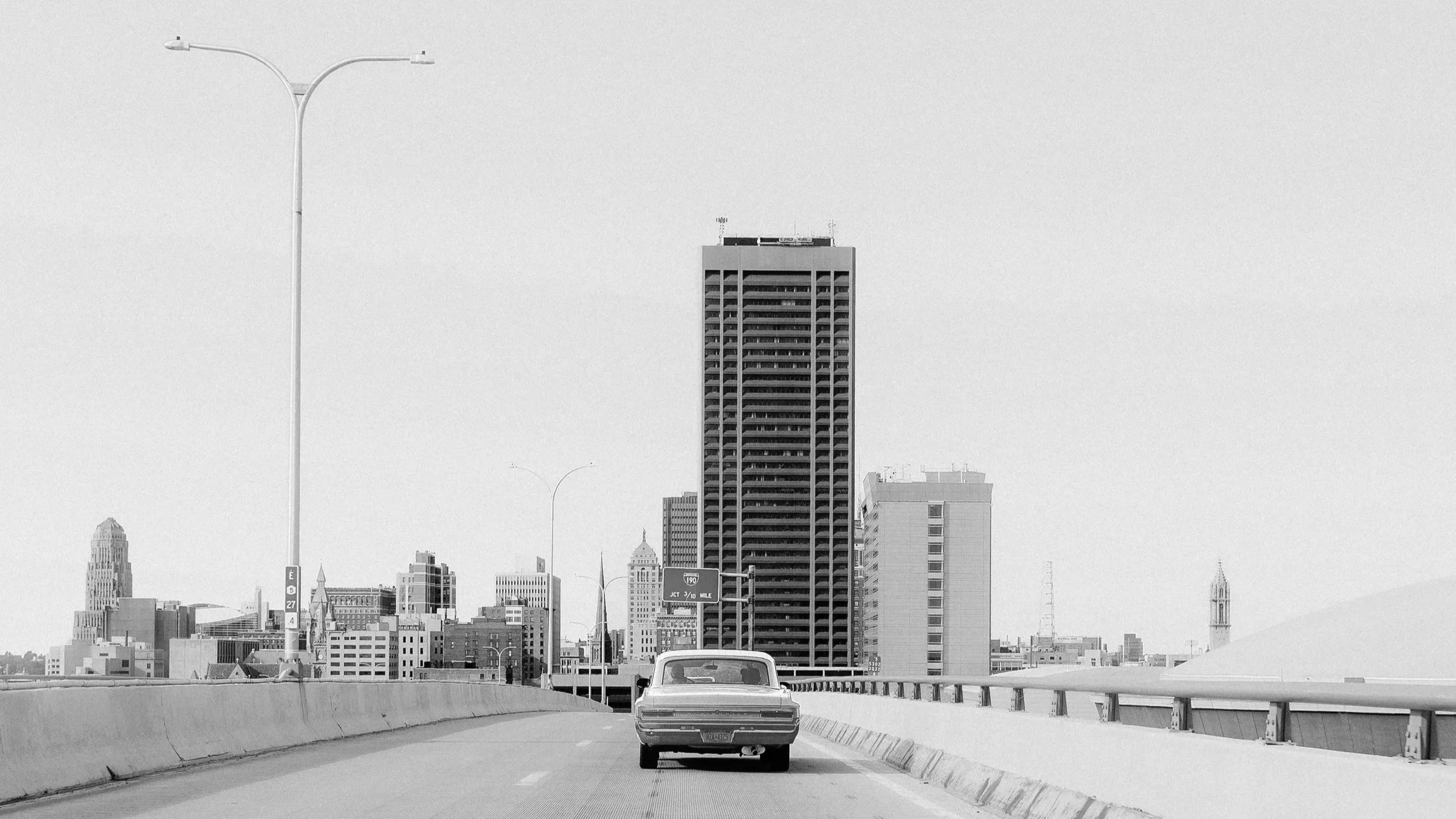 Black and white photo of a city skyline with a tall building, other skyscrapers, and a church steeple in the distance, taken from a highway with a car driving away from the camera.