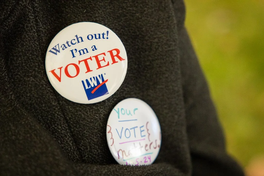 Close-up of two buttons on a dark jacket, one stating "Watch out! I'm a VOTER LWV" and the other with handwritten text about voting.