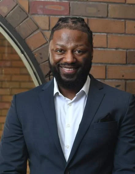 A man with dark skin and braided hair in a bun, wearing a navy blue suit and white shirt, smiling in front of a brick wall.