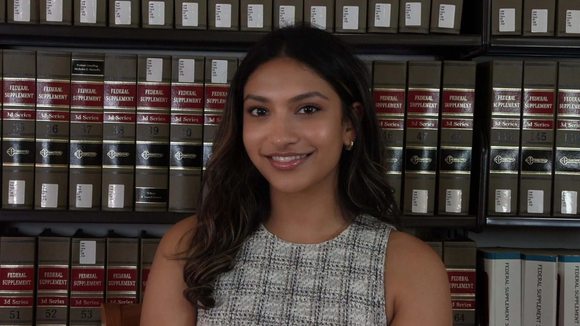 Young woman with long wavy hair and gold earrings smiling in front of bookshelves filled with legal books and federal supplement volumes.