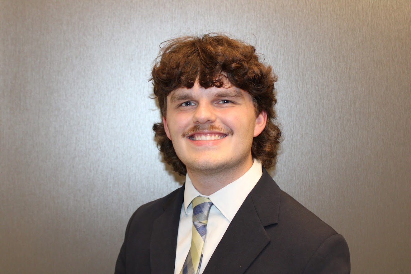 A smiling young man with curly brown hair, wearing a black suit, white shirt, and patterned tie, standing in front of a plain gray wall.