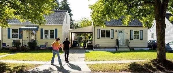 Two people walking on a sidewalk in front of two houses with trees and green grass.