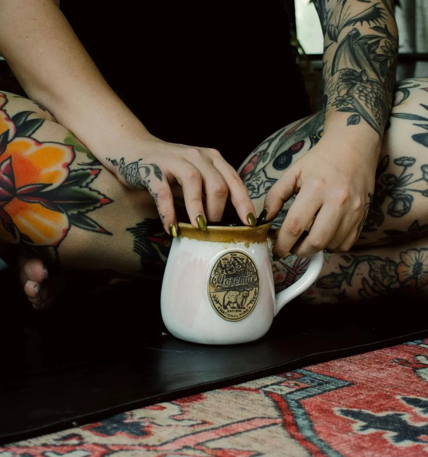 Person with colorful tattoos on arms and hands, holding a Yosemite National Park branded mug with brown coffee or tea, sitting on a floral patterned cushion or blanket | Yoga Classes and Events | San Antonio, TX