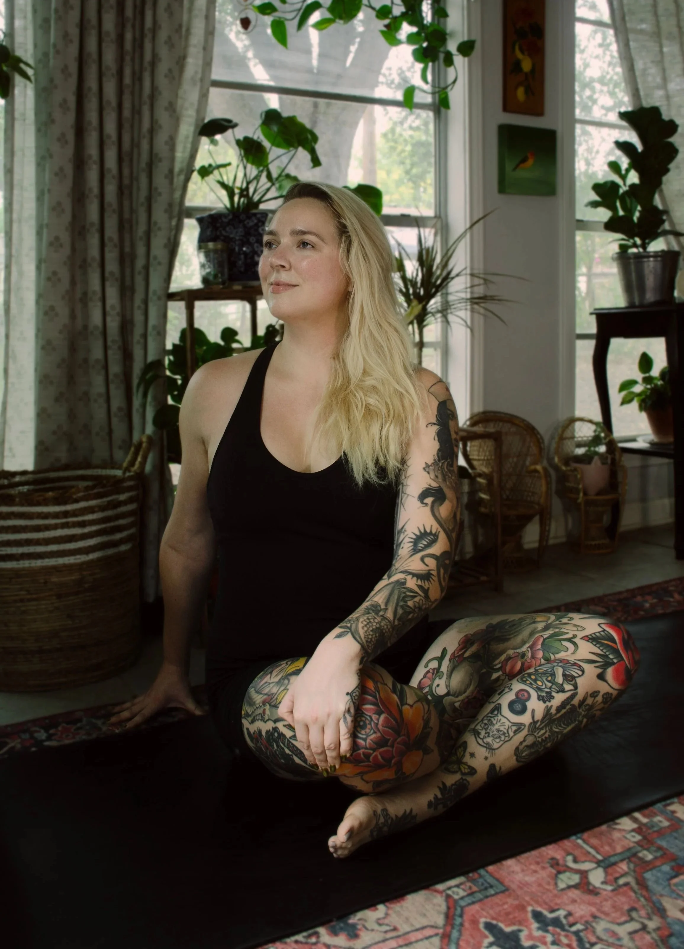 A woman with long blonde hair, tattoos on her right arm and legs, sitting on a black mat in a room filled with natural light, surrounded by plants, paintings, and wicker furniture | Yoga Classes and Events | San Antonio, TX
