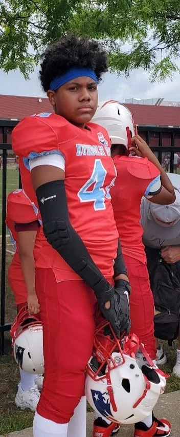 A young football player with curly hair, wearing a red Humboldt Park Patriots jersey with the number 44, standing outdoors on a football field during daytime, holding a white helmet with a red and blue star logo, and a serious expression.