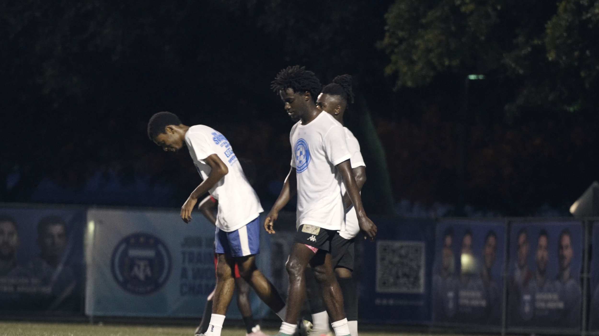 Soccer players walking on the field during a night game, wearing white shirts and shorts.
