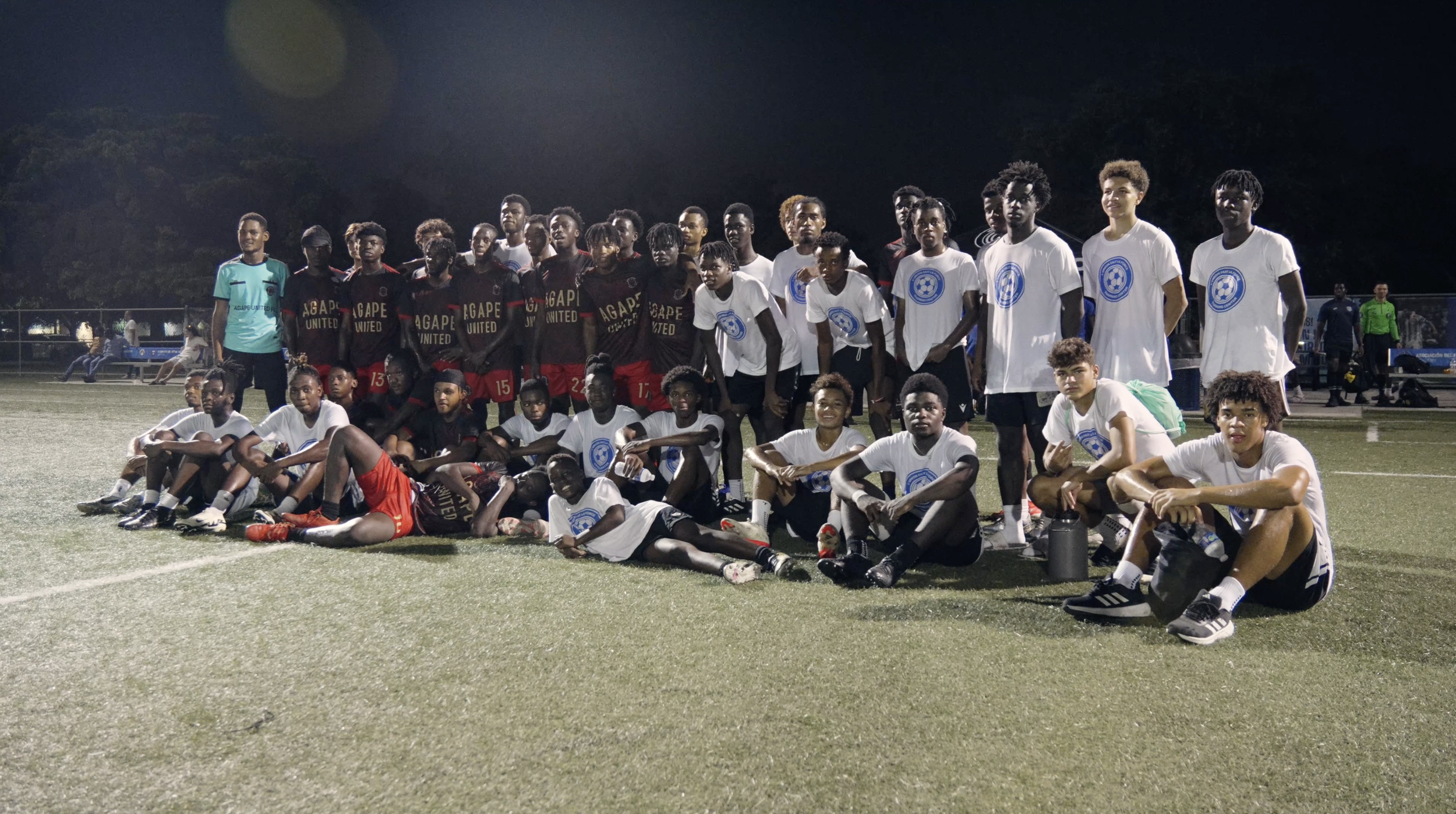 Group photo of a youth soccer team on a field at night, some players in white shirts with blue logos, others in black and red uniforms, with a coach in a green shirt on the left.