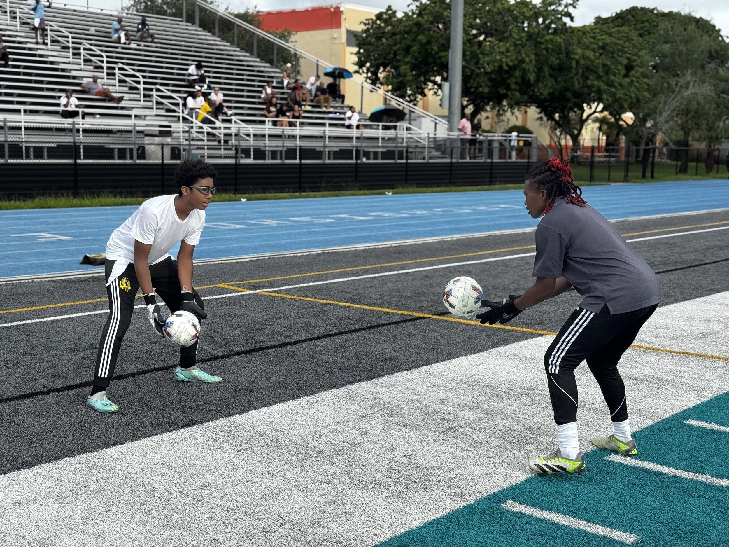 Two young male soccer players practicing on a sports field, holding soccer balls in preparation for a drill, with spectators sitting on bleachers in the background.