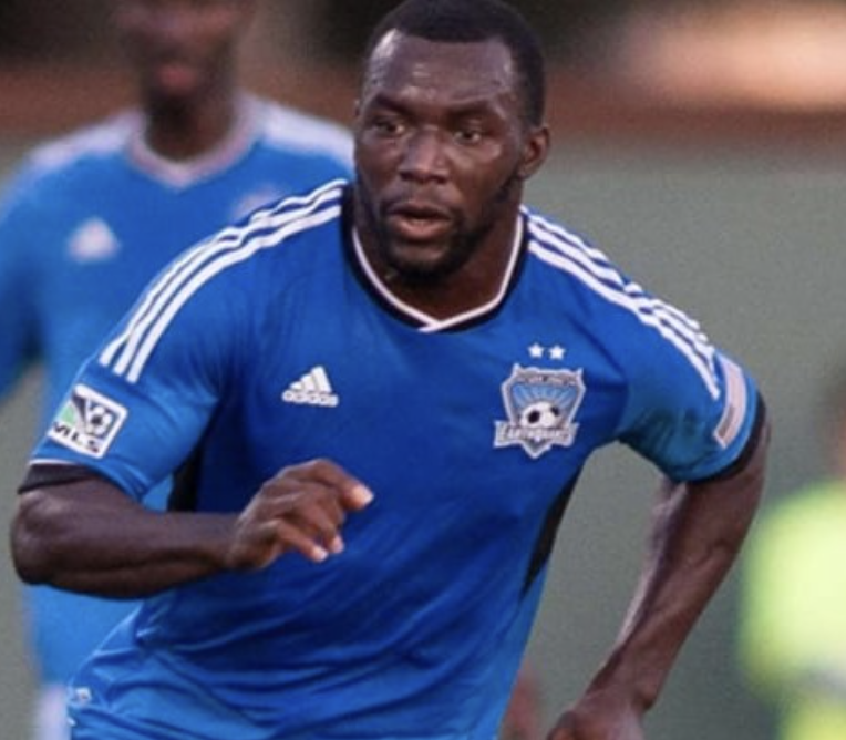 A male soccer player with dark skin wearing a blue jersey with white stripes, running during a match.