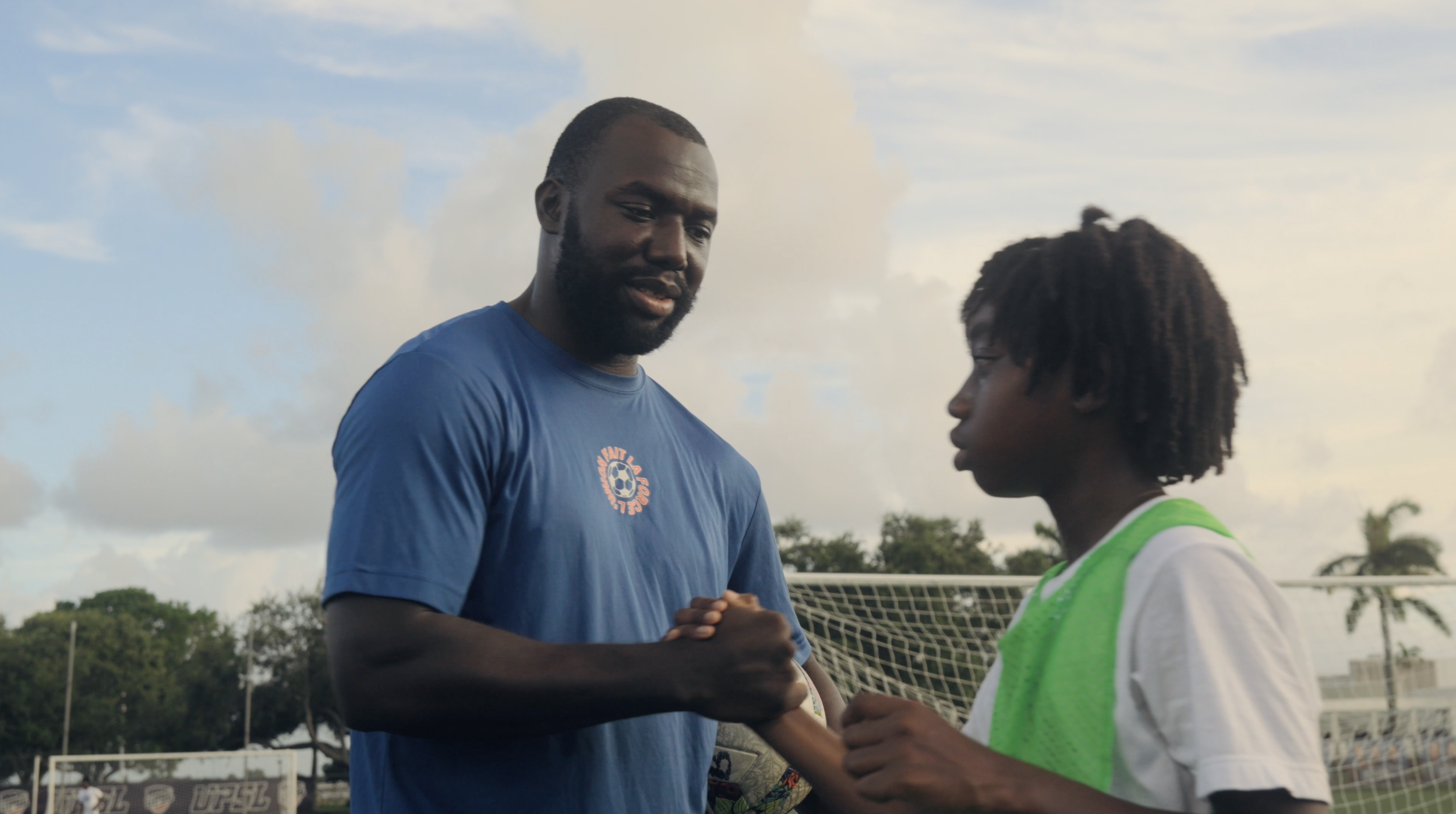 A soccer coach and a young player shake hands on a soccer field during daytime, with goalposts and trees in the background.
