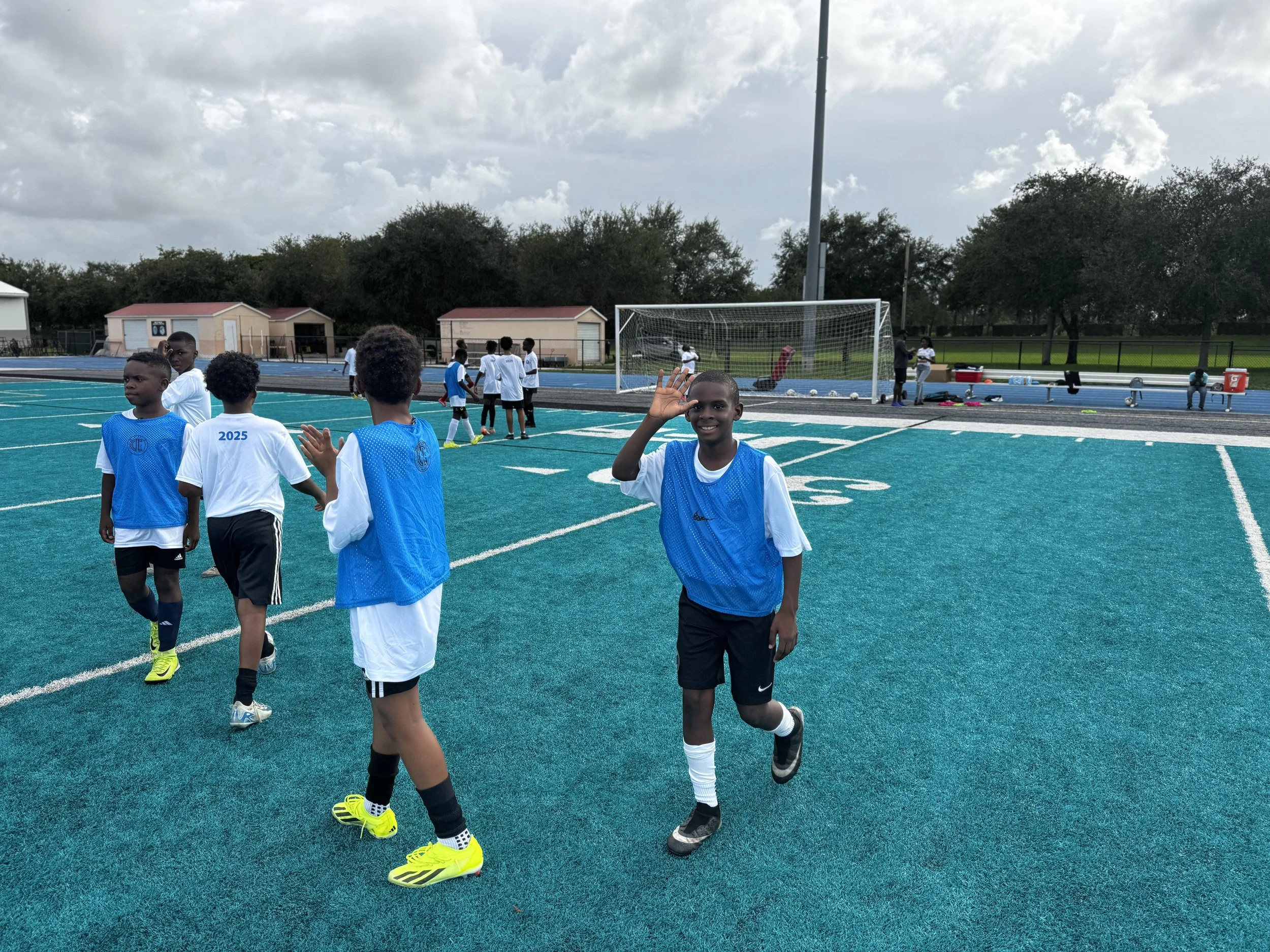 Young boys in sports uniforms on a blue turf field, some warming up and others walking towards the goal, with a cloudy sky overhead.