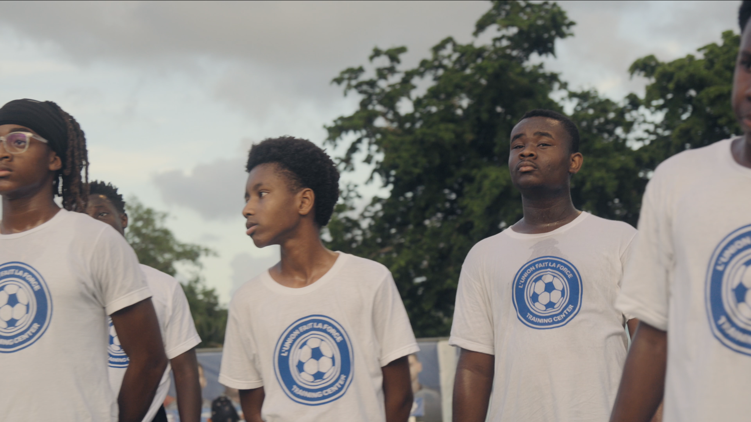 Group of young people standing outdoors wearing white T-shirts with a blue and white soccer emblem, some with serious expressions, with trees and cloudy sky in the background.
