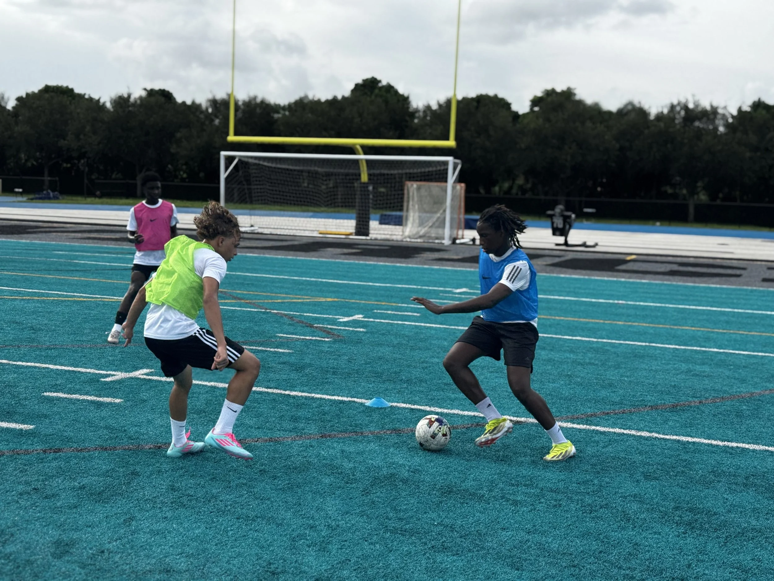 Three kids playing soccer on a blue outdoor field, with one wearing a blue vest and two wearing pink and green vests. A goal and track are visible in the background.