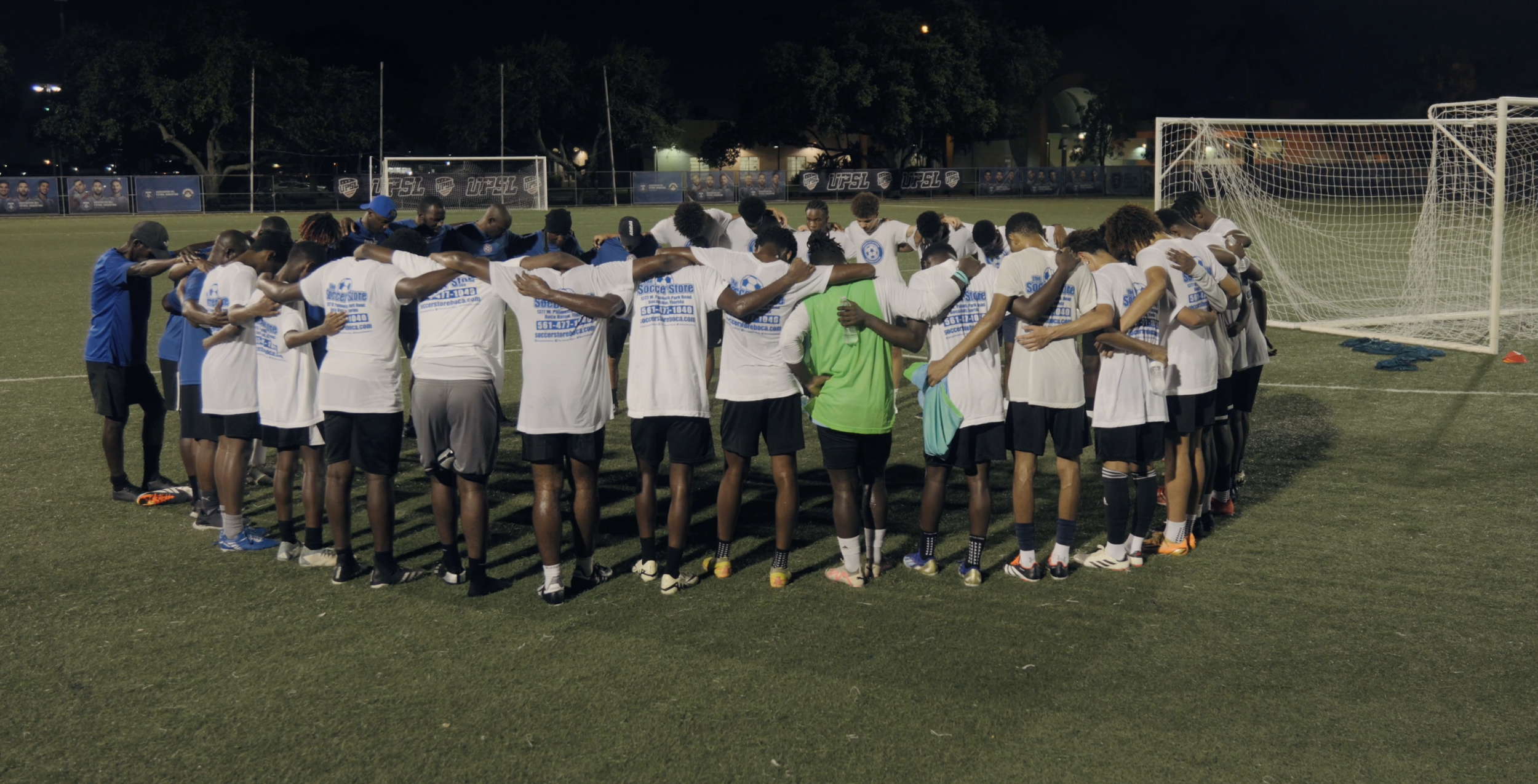 Group of soccer players in white and blue jerseys standing in a circle with their arms around each other's shoulders on a soccer field at night, possibly before a match or practice.