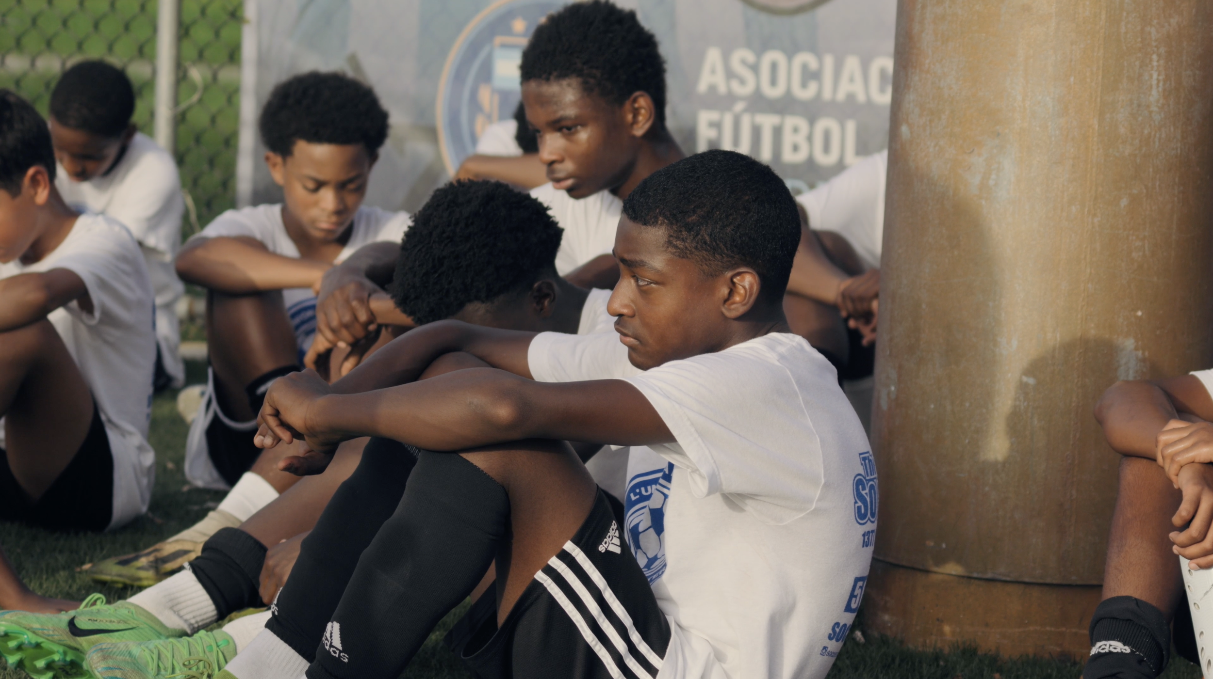 Group of young male soccer players sitting on the ground during a break, wearing sports uniforms, with a soccer field and a banner in the background.