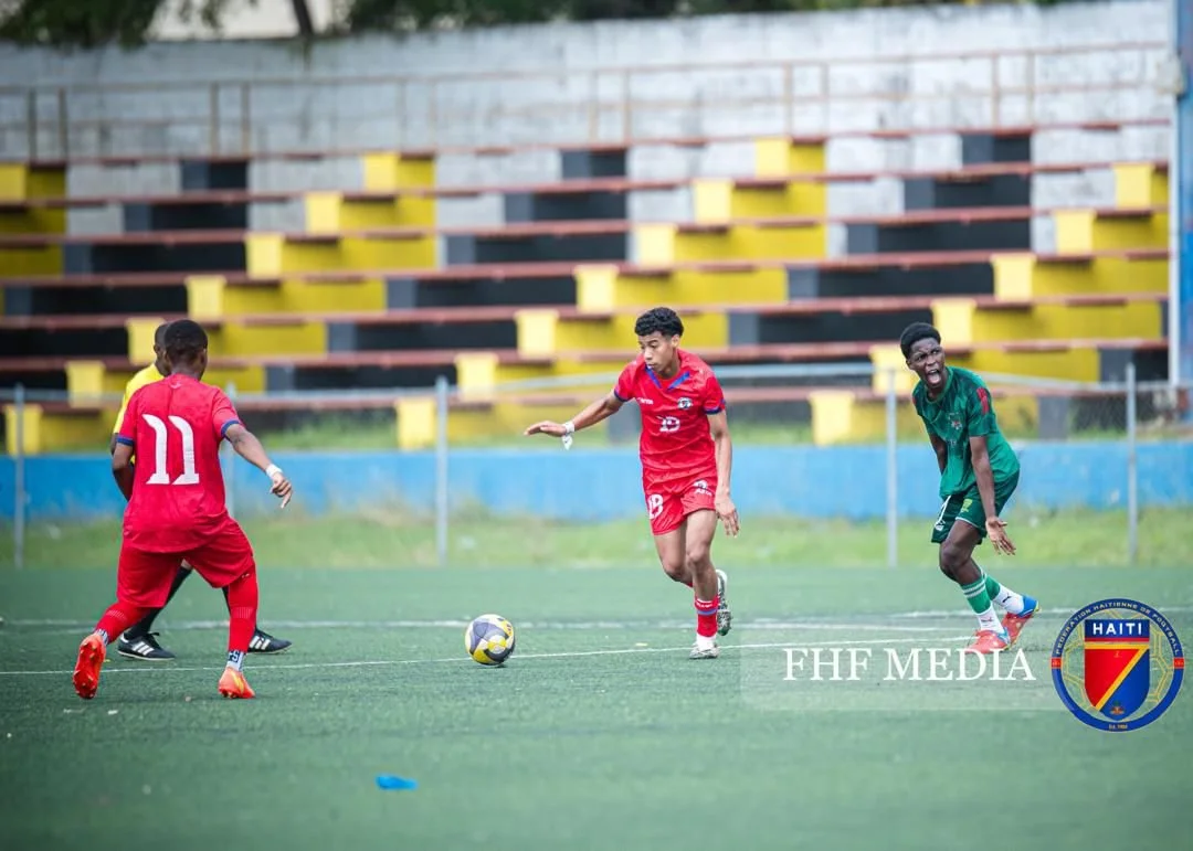 Three young male soccer players on a green field, two in red uniforms and one in green, engaging in a game with a black and white soccer ball. Empty bleachers with yellow and black seating are visible in the background, and the players appear focused