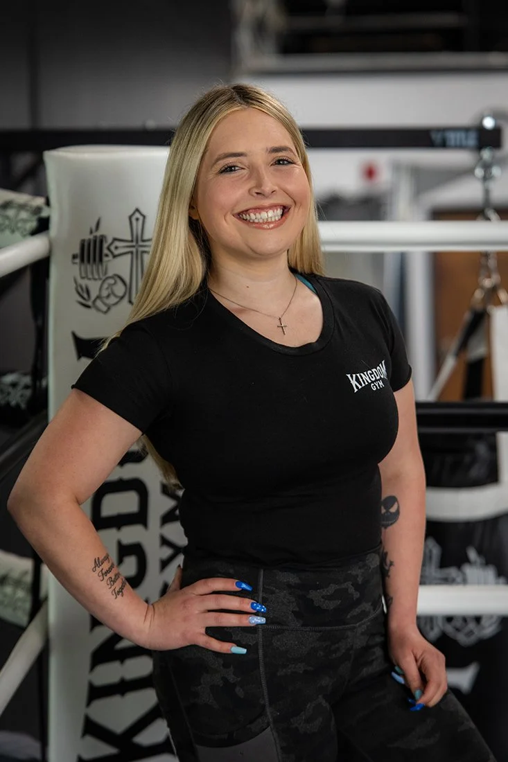A woman with long blonde hair smiling, wearing a black t-shirt with white text that says 'Kingdom Gym', standing in a gym with workout equipment in the background.