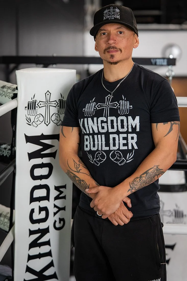 Man wearing a black 'Kingdom Builder' t-shirt and a black cap with the word 'Kingdom Gym,' standing in a gym next to a white gym equipment stand with the World's Gym logo.
