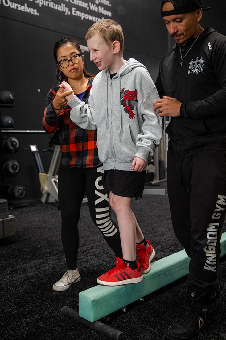 A young boy in a gray hoodie and black shorts is balancing on a foam roller, with a fitness trainer and a woman supporting him in a gym. The trainer is wearing black athletic gear with 'Kingdom Gym' written on it, and the woman is wearing a red and black checkered shirt. They are in a gym with black walls and fitness equipment in the background.