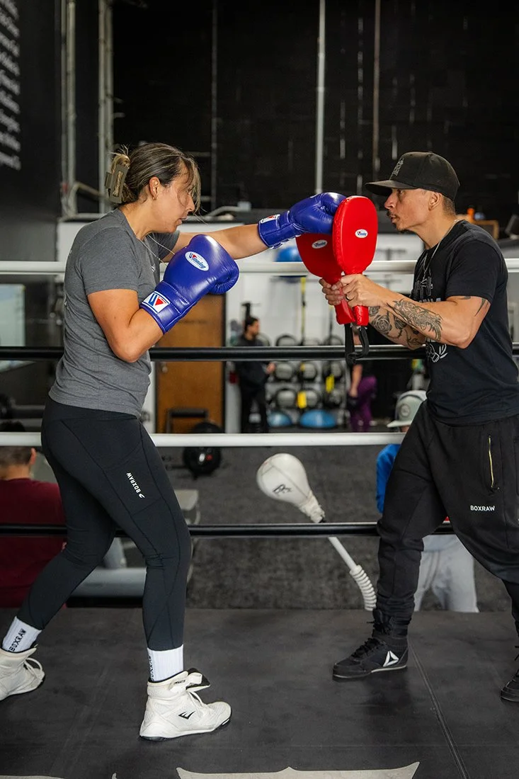 Woman sparring with a man in a boxing gym, she is wearing blue boxing gloves and performing a punch on a red focus mitt held by the man, who is holding a red focus mitts in front of him.