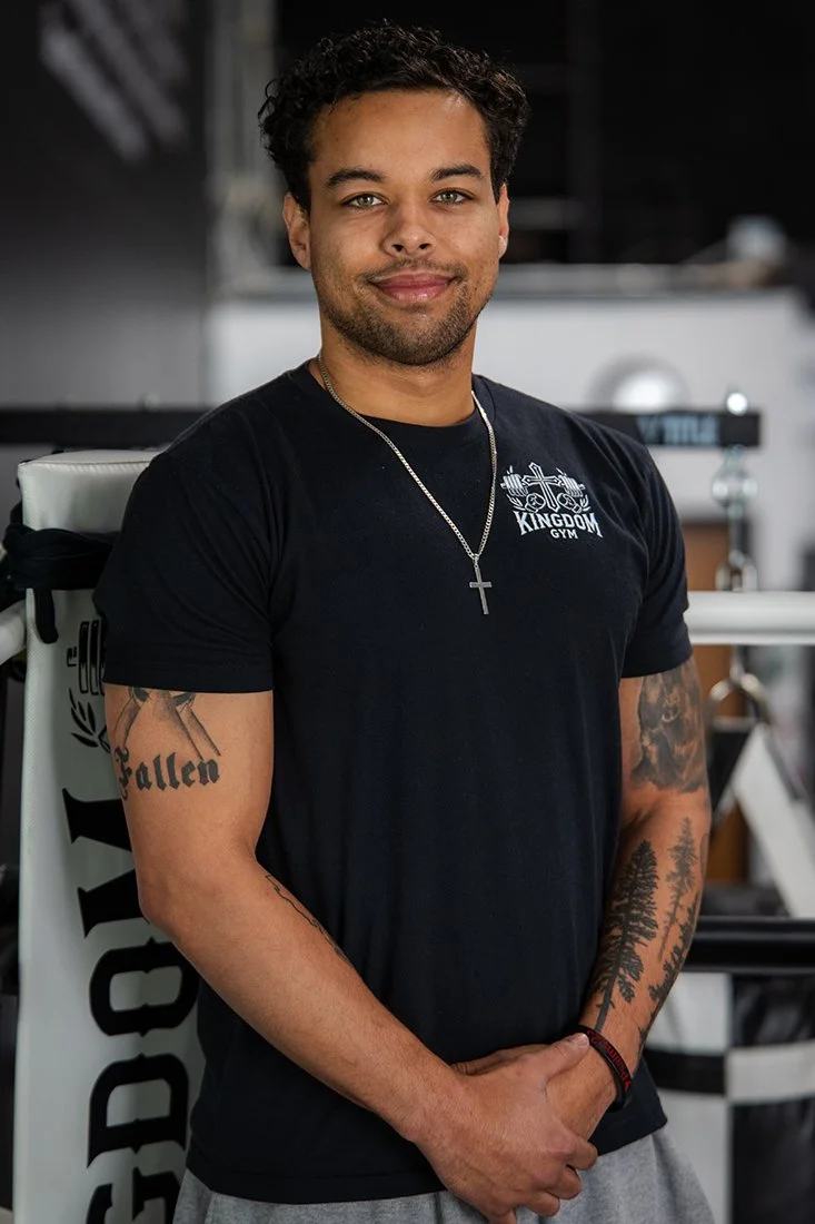 A young man with short curly hair, wearing a black T-shirt with 'Kingdom Gym' logo, a cross necklace, and tattoos on his arms, standing inside a gym.
