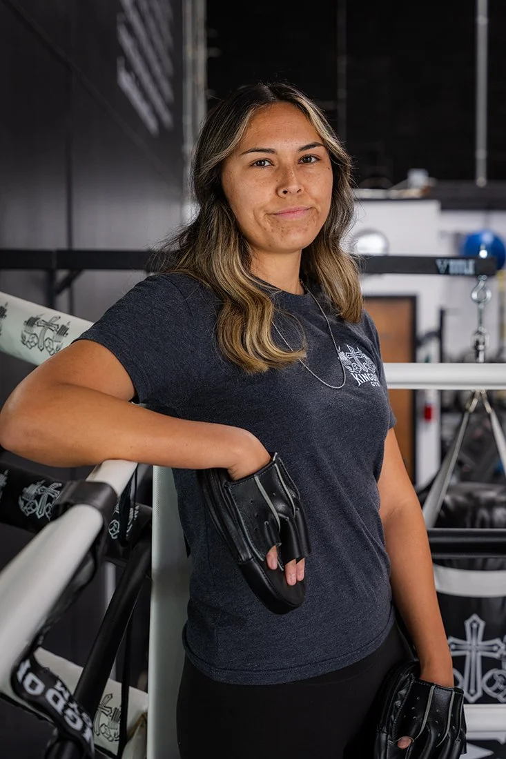 A young woman in a black t-shirt and workout gloves standing in a gym with her arm resting on a rowing machine.