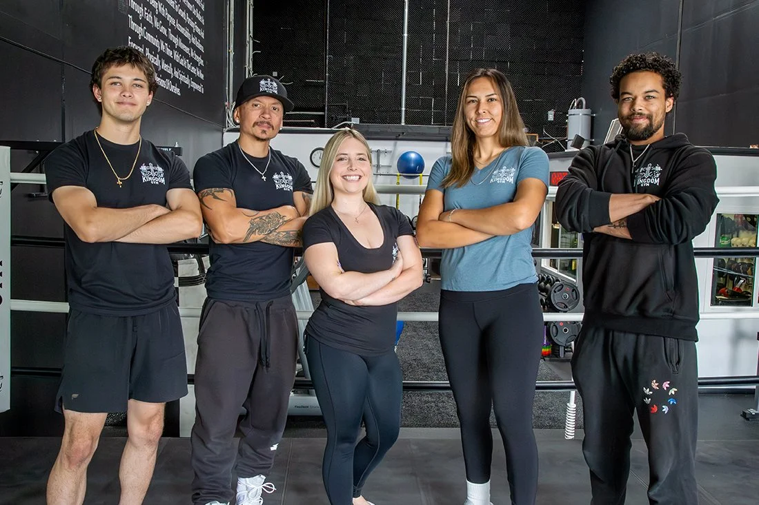 Five fitness trainers standing with arms crossed inside a gym, smiling at the camera.