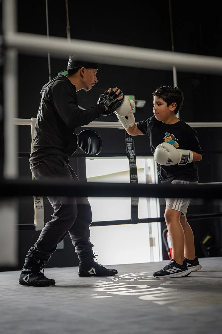 A boxing coach coaching a young boy in a boxing ring, both wearing boxing gloves, with the coach demonstrating a technique.