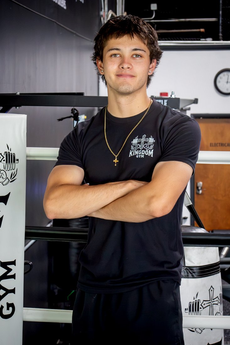 Young man with dark hair and light skin standing in a gym, crossing his arms, wearing a black T-shirt with the words "Kingdom Gym" and a cross logo on it, and a gold chain with a cross pendant.