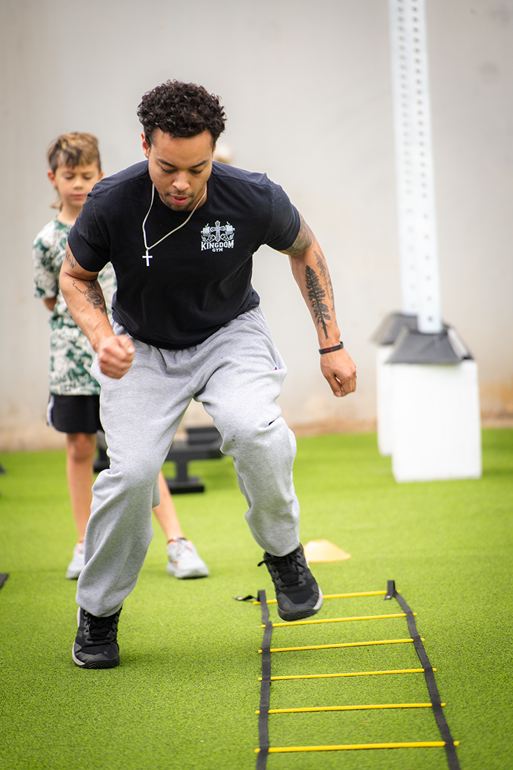 A man with tattoos wearing a black t-shirt and gray sweatpants training with agility ladder in an indoor gym, with a boy in the background.