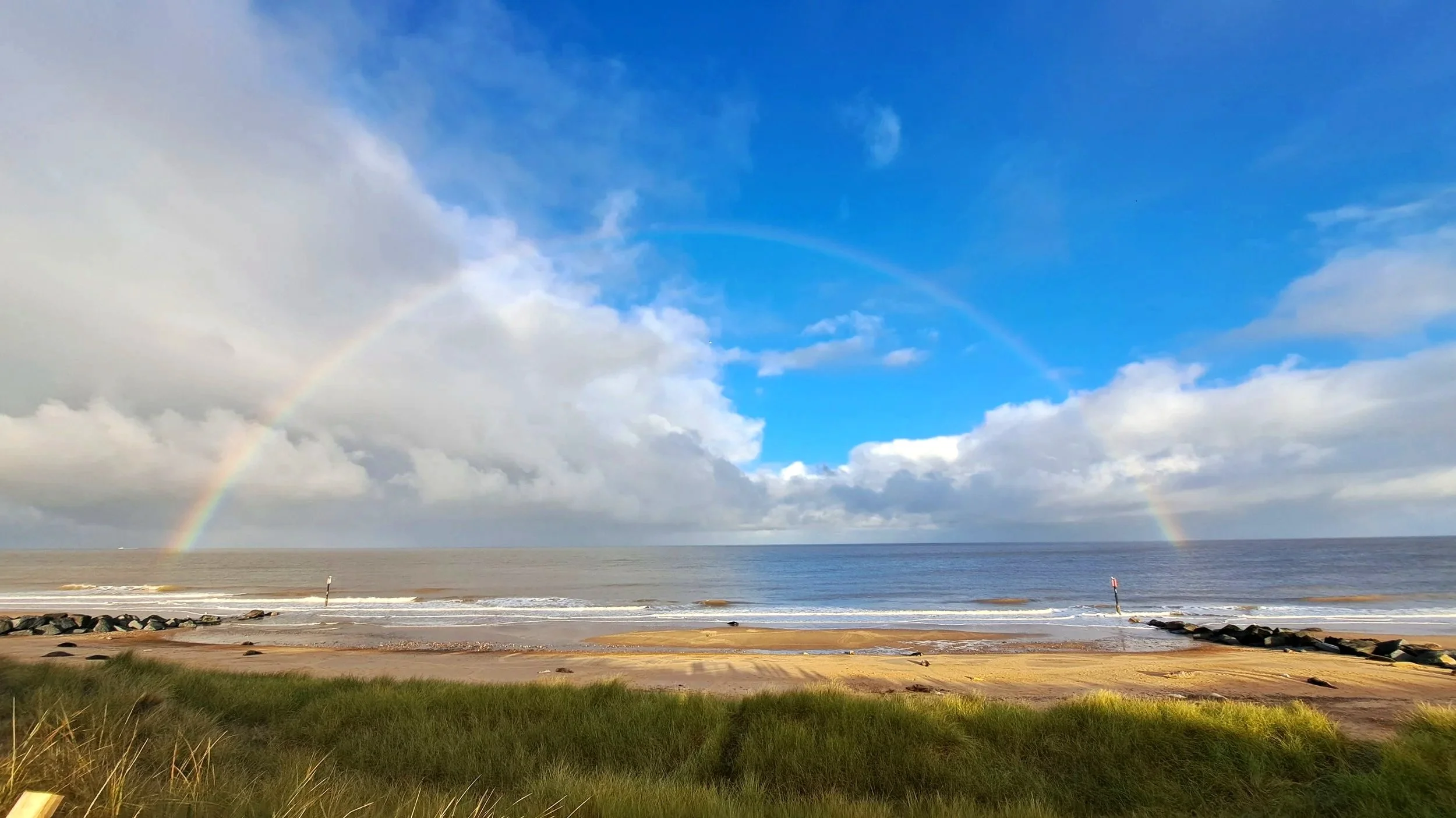 A beach with grassy dunes in the foreground, a sandy shore with rocks, and the ocean in the background. A rainbow arches across a partly cloudy sky over the water.