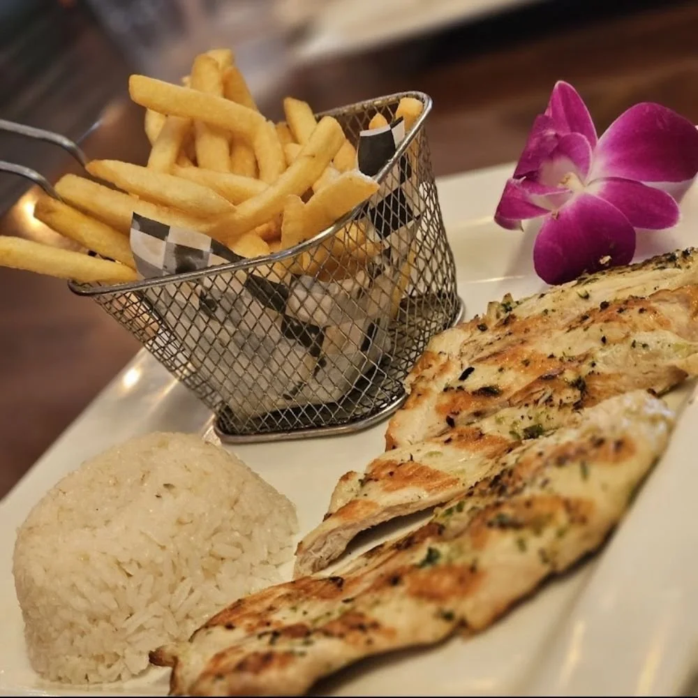 Grilled chicken breast with herbs, steamed rice, a side of French fries in a small metal basket, and a pink orchid flower on a white plate.