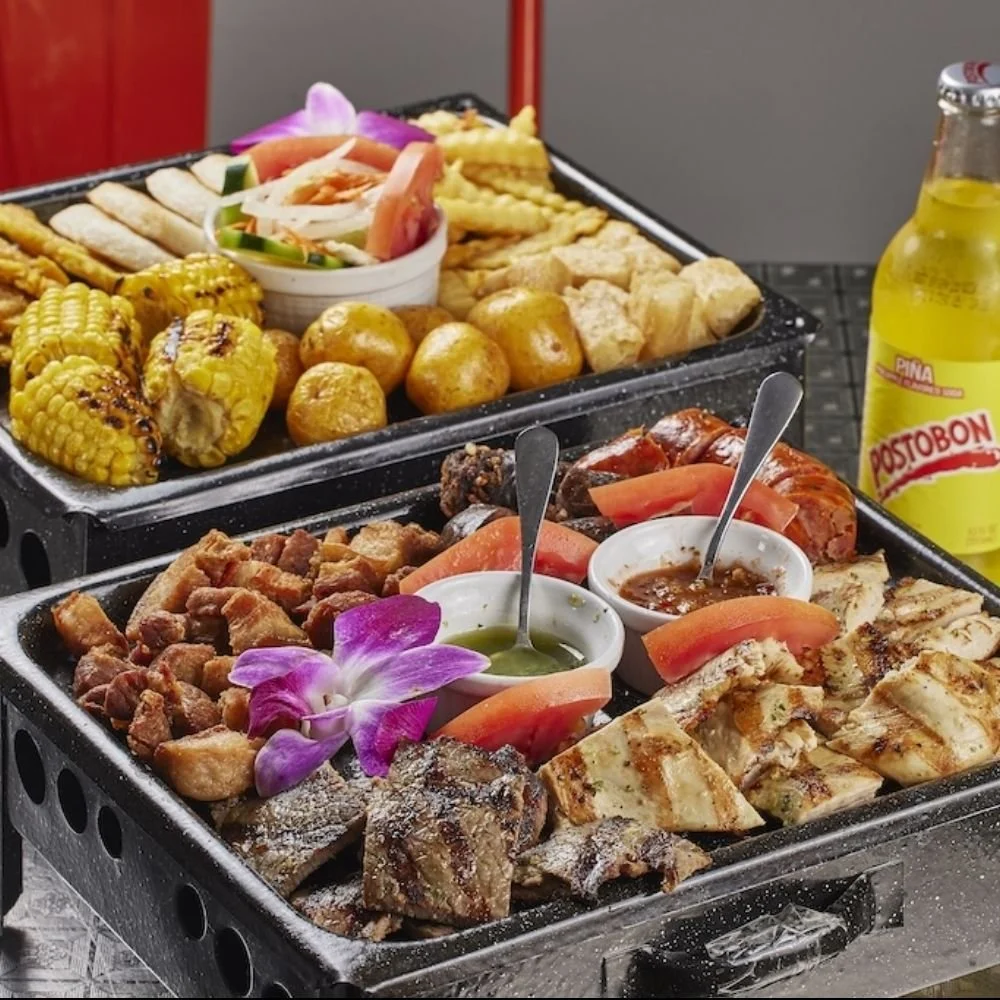 Two black trays with grilled corn, fried balls, fried plantains, and salad, with two small bowls of dipping sauces, garnished with purple orchids, and a bottle of Postobon lemon soda in the background.