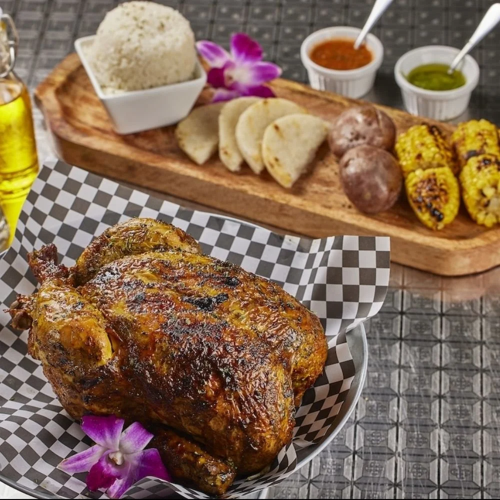 Roasted chicken served with grilled corn, potatoes, and bread, with rice and sauces on a wooden platter in the background.