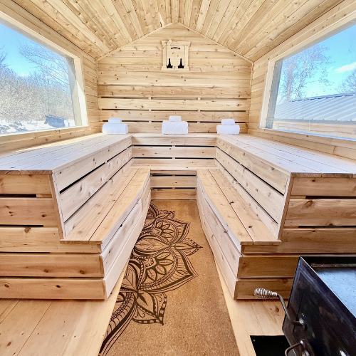 Wooden sauna room with bench seating, large windows on both sides, and a decorative rug on the floor.
