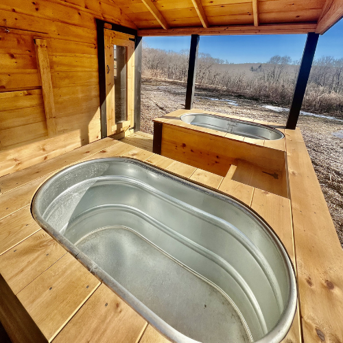 Two metallic bathtubs inside a wooden shelter overlooking a scenic outdoor landscape with trees and clear blue sky.