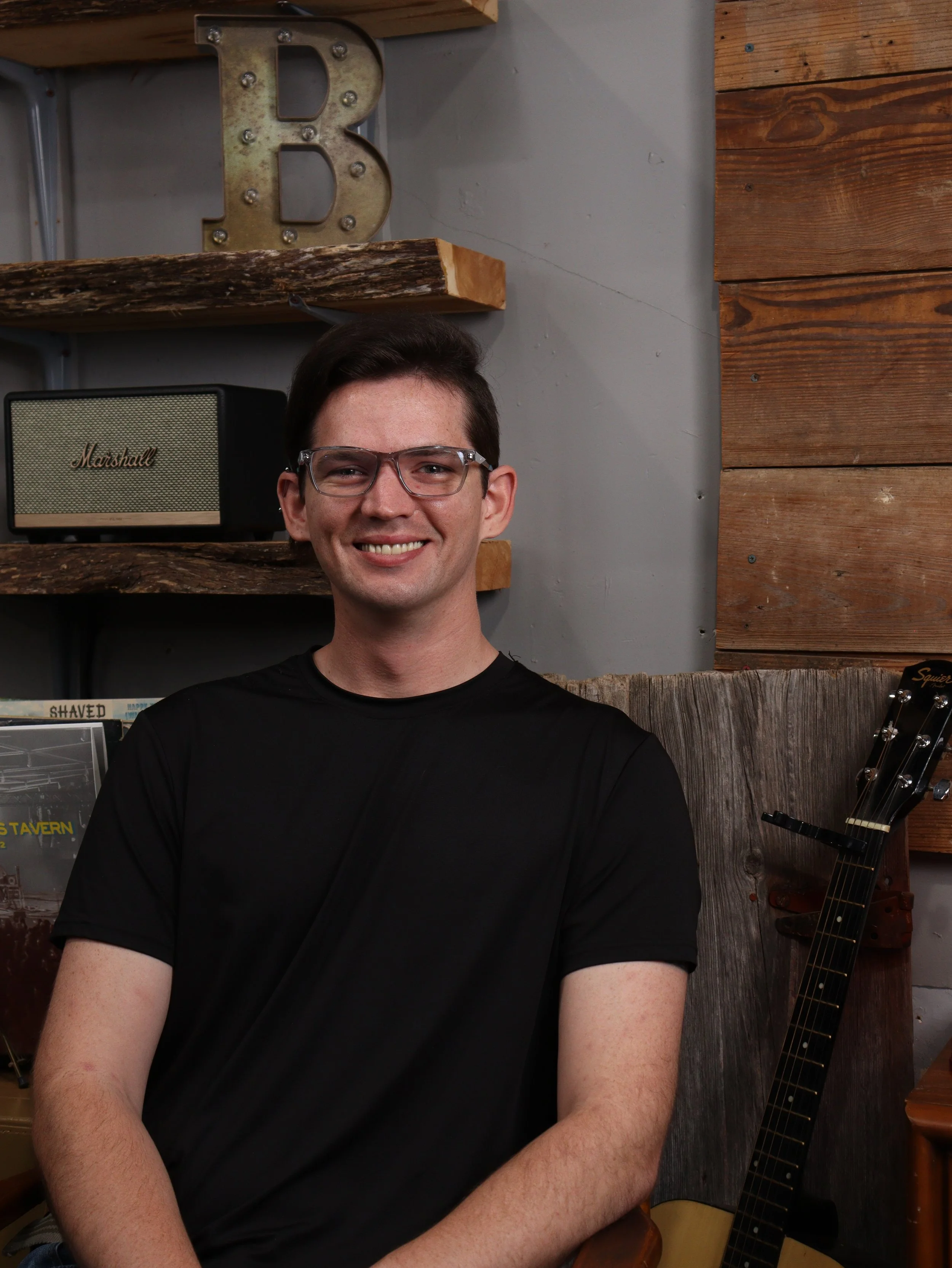 A man with short dark hair, glasses, and a black shirt smiling, sitting in a rustic indoor setting with wooden shelves, a decorative metal letter B, and a guitar in the background.