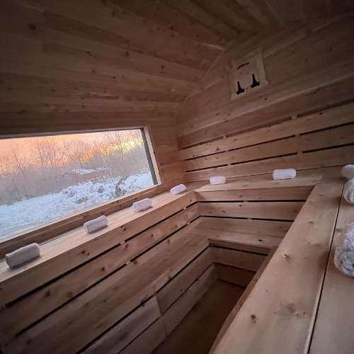 Interior of a wooden sauna with a bench, a large window showing snowy landscape outside, and some sauna stones placed along the window ledge.