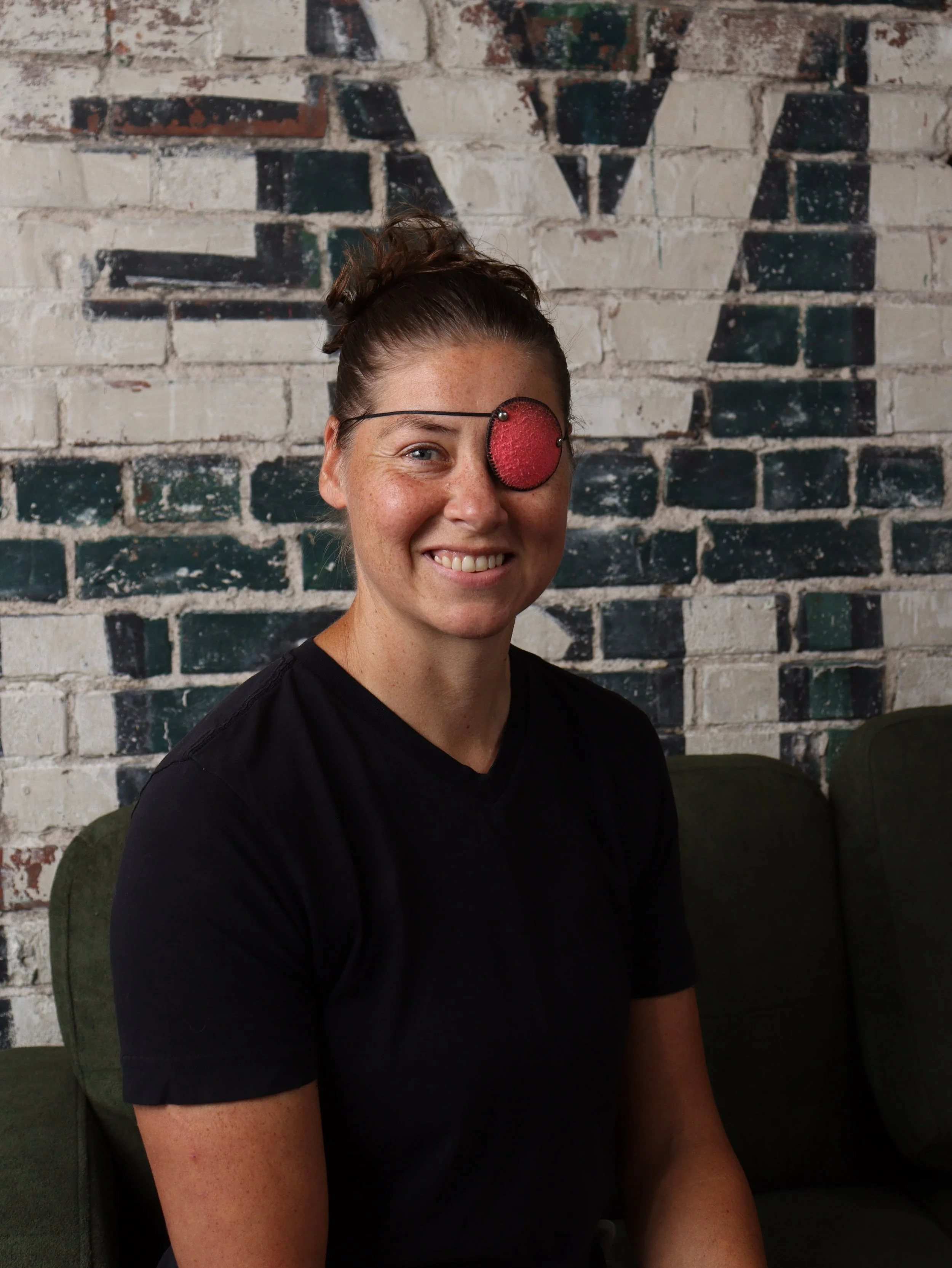 A woman with light skin, freckles, and brown hair tied in a bun, smiling and wearing a black shirt. She has a red eye patch over her left eye and is sitting against a brick wall with graffiti.