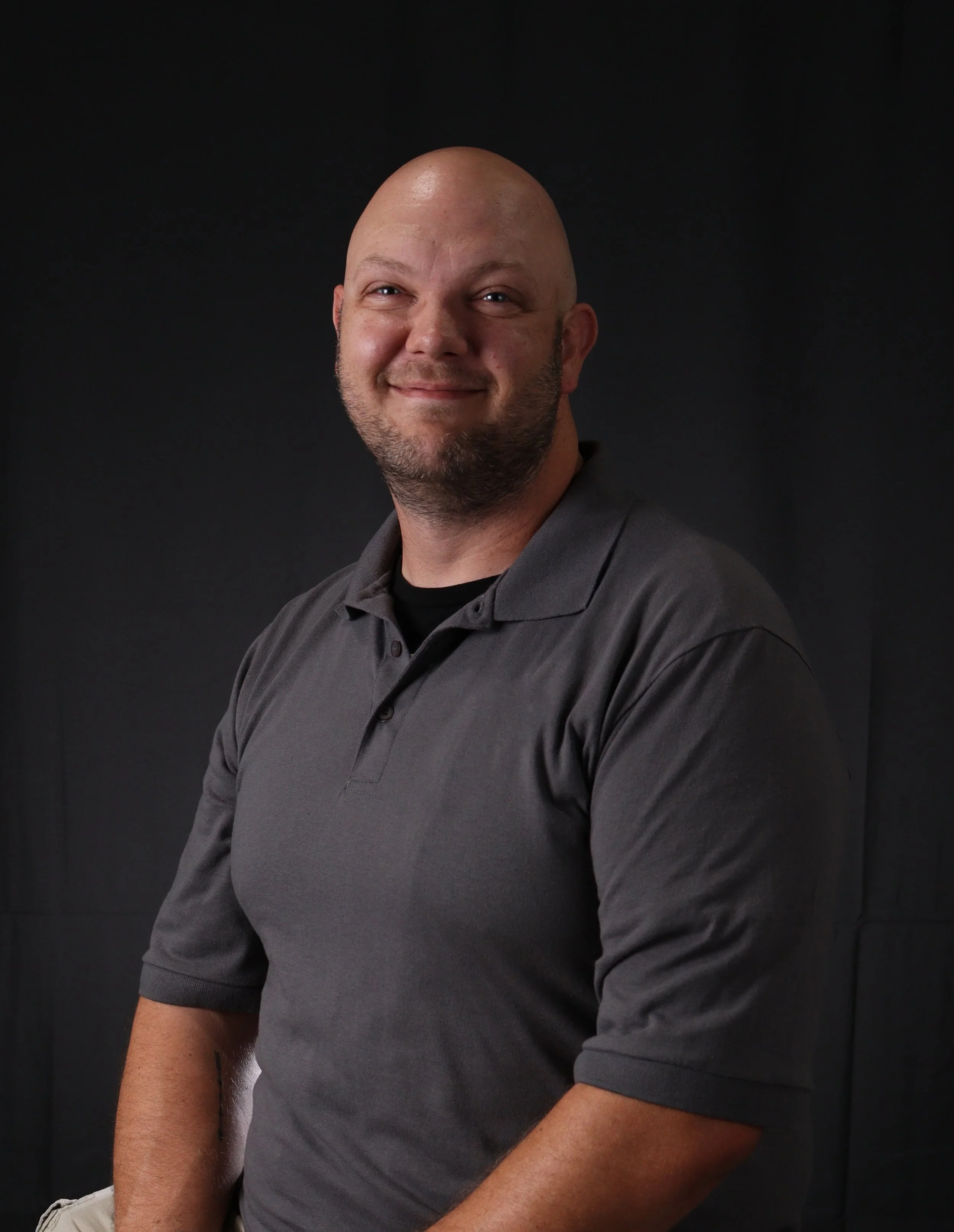 A smiling man with a bald head and beard, wearing a gray polo shirt, posed against a dark background.